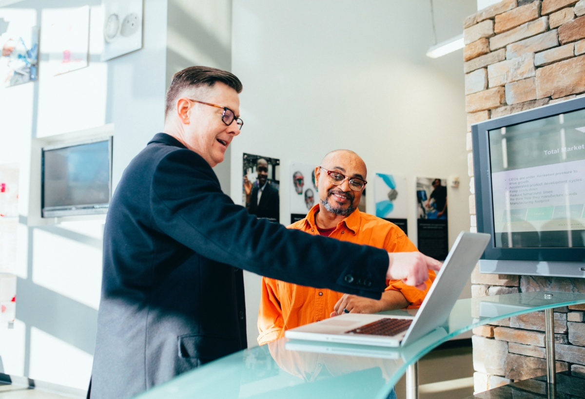 Man pointing at a grey laptop and smiling with another man watching him and smiling